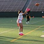 Two women's flag football teams play a match on an athletic field. A ref moves in to watch the play