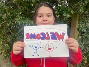 A middle-school age student poses for a photo with a hand drawn sign that says Welcome in English