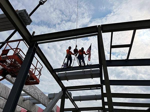 two people standing on a steel beam as another is raised toward them with a flag on it