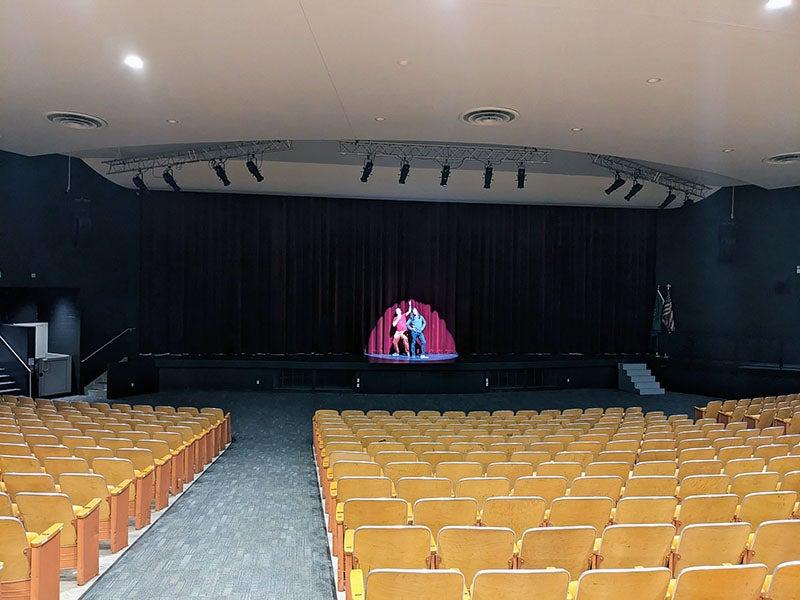 an auditorium with light brown seats looks toward a stage where two people are in the spotlight