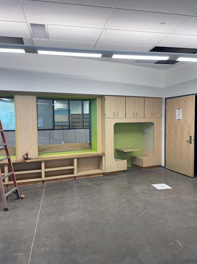 a classrom wall section has a nook below cabinets that includes two facing benches with a ledge between them. next to them is open shelving below a window. Both areas are highlighted by bright green accents