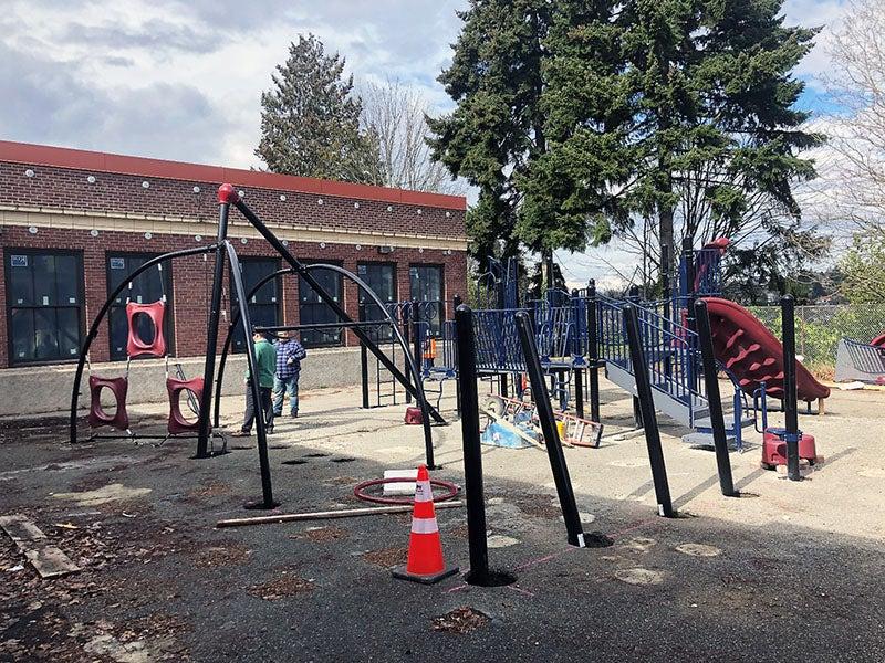 outside of a 1-story brick building, playground equipment is being installed. there are trees in the background