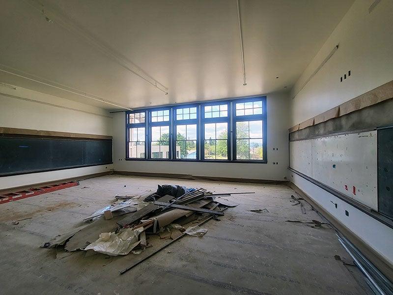 a classroom has white boards on the left wall, windows on the far wall, and a blackboard on the right wall. the floor is covered with construction protection and a pile of material is in the middle of the floor