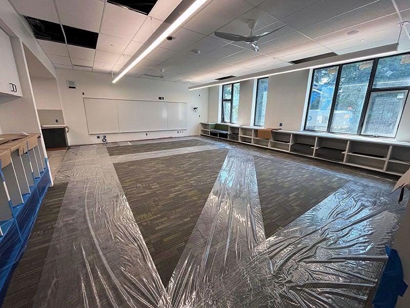 a classroom has shelves under the windows, carpet with plastic runners, and a white board