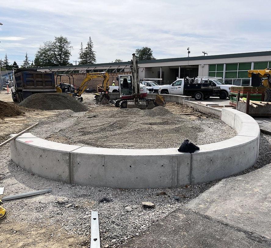 a semi-circular low wall has gravel piled in the center; construction equipment and a building are behind it