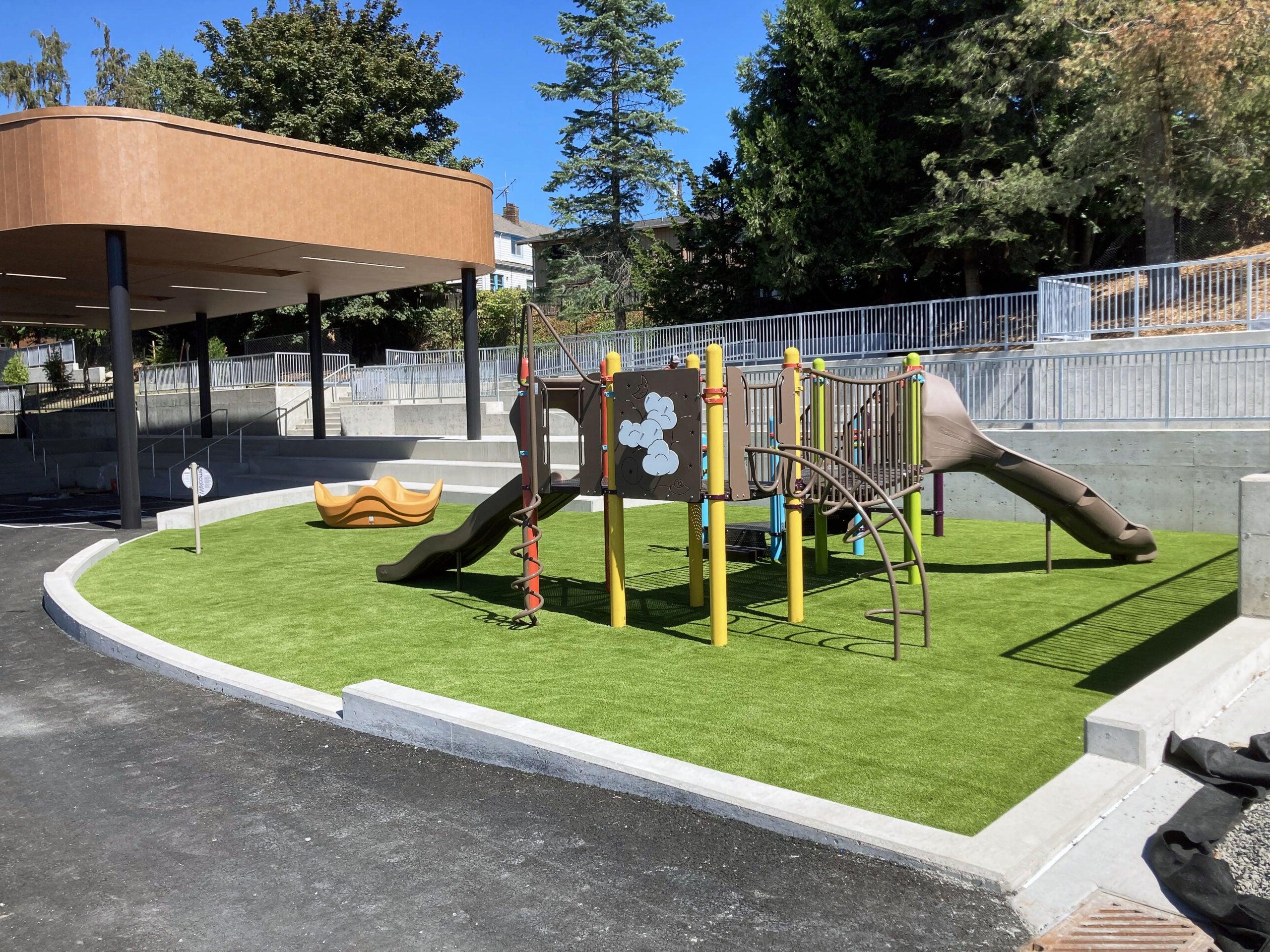 playground equipment sites on a green surfaced area with curbing around it. a building is behind it