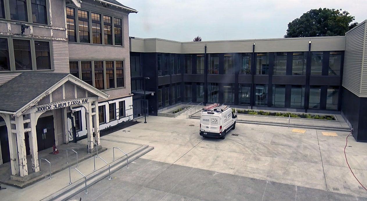 a three story historic building sits to the right with a new 2 story building directly in center with paved area in front of both