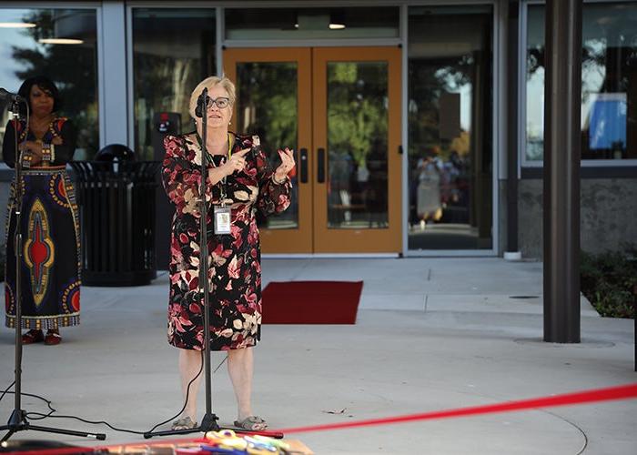 a woman in a maroon dress with pink flowers stands in front a microphone behind a red ribbon