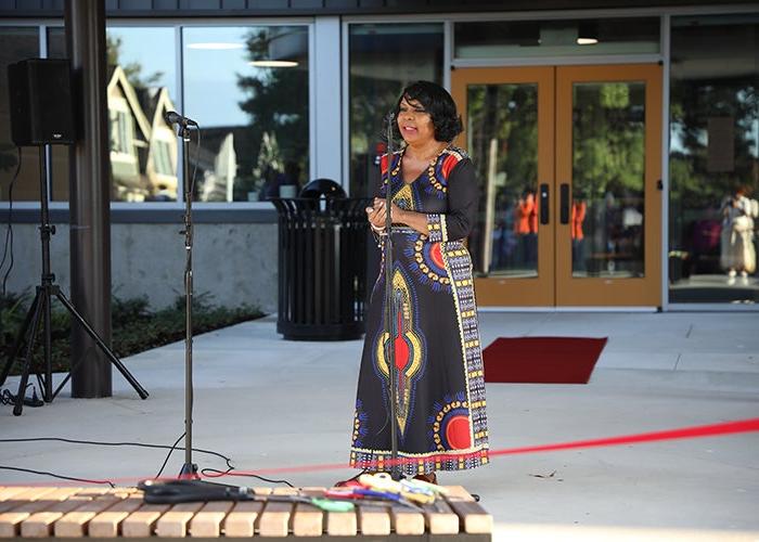 a woman in a long blue dress with red and gold figures stands in front of a microphone behind a red ribbon