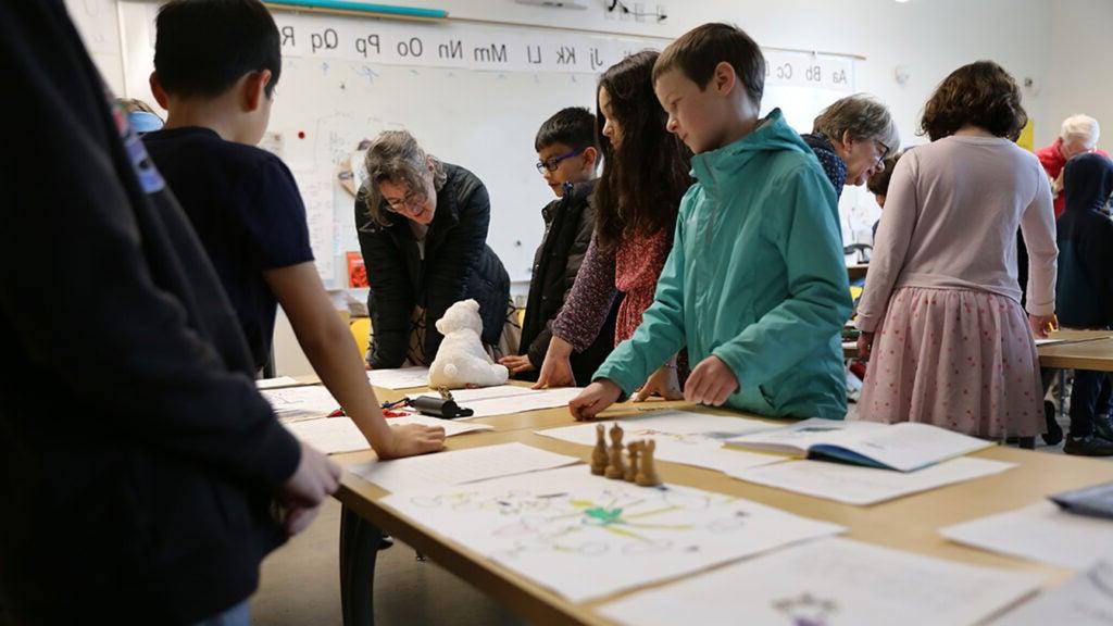 Students and elders stand around desks covered in artifacts, discuss objects.