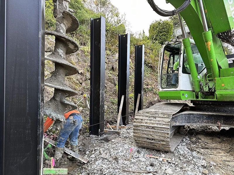 a large construction machine with tracks has a giant drill bit and is screwing it into the ground between two metal posts