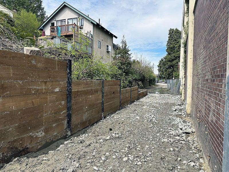 a brick wall on the left faces a partial wall made of steel posts and wood slabs on the right. a house is visible on the hill behind the right side wall