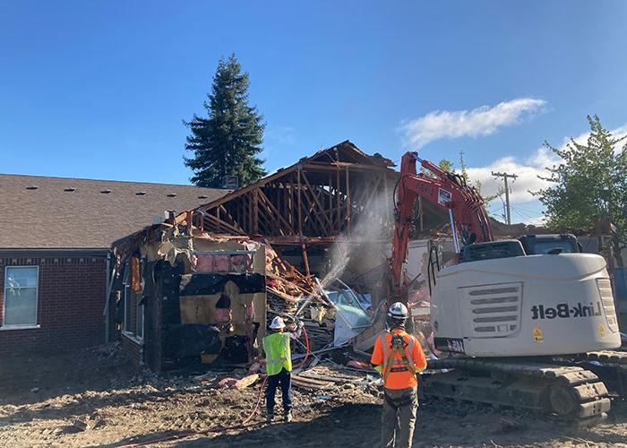 a bucket loader is demolishing a portion of a brick building while workers spray water