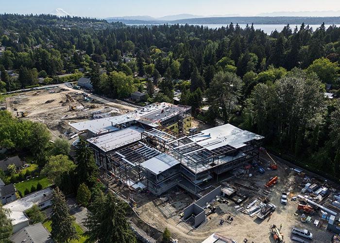 aerial view of a construction project with a large H-shaped building in progress. Behind it is a open dirt area with construction equipment. Trees are all around and a lake is visible in the far background.