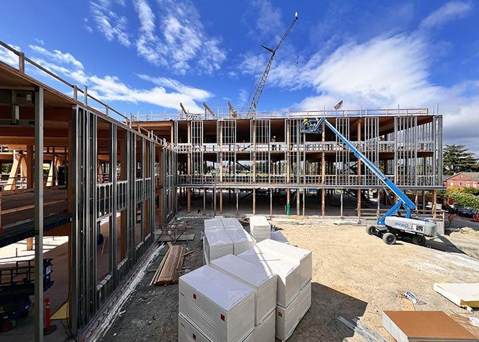 exterior of a large building under construction with walls partially framed, a person on a lift near the 3rd floor, construction materials in foreground and bright blue sky with light clouds.