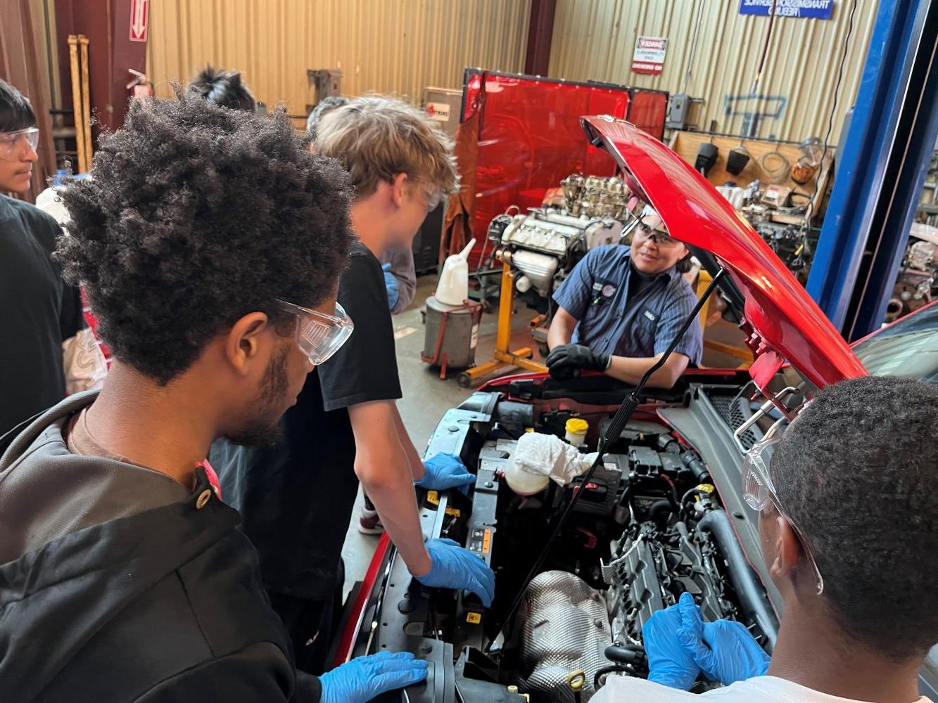 Three students looking at car with hood popped and teacher