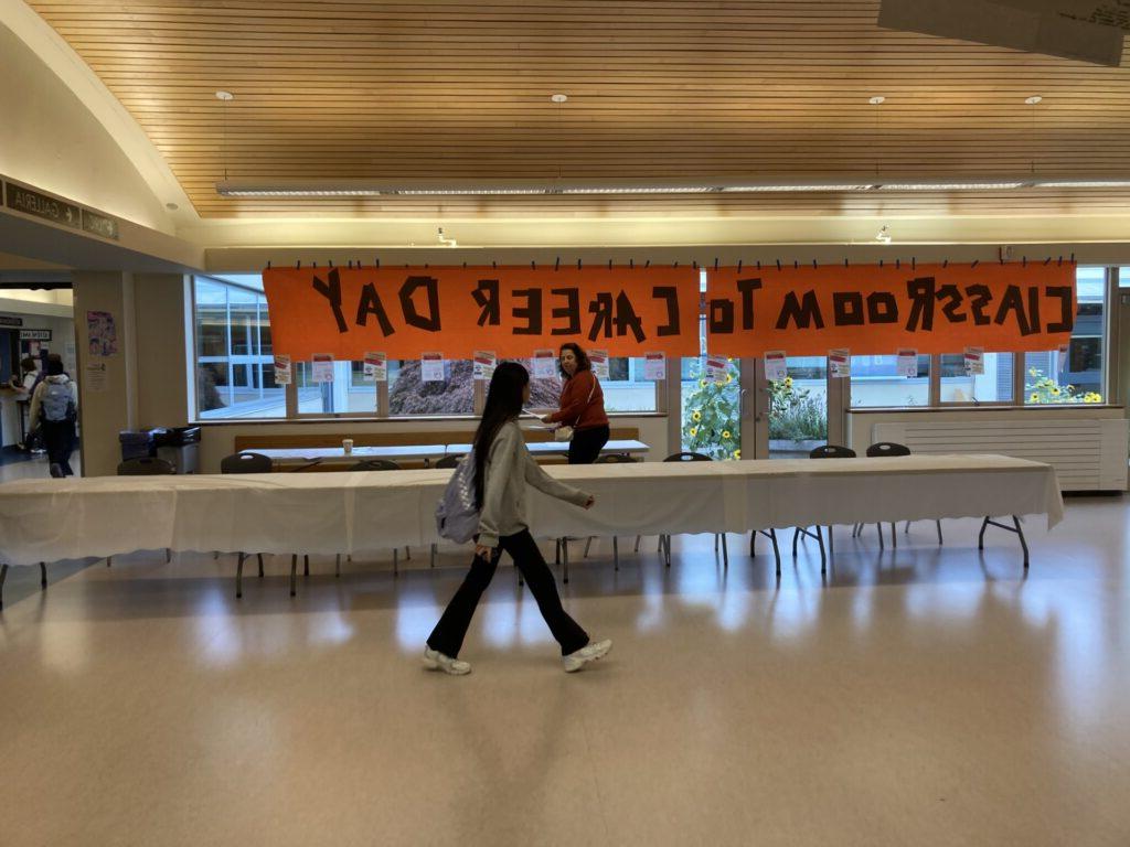 Student walking in front of set up table in front of large orange banner with words "classroom to career day" written on it. 